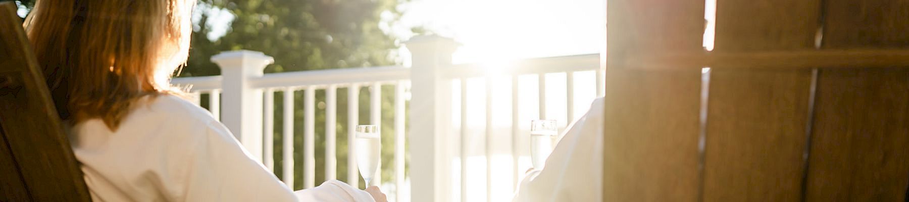 Two people sit on wooden chairs on a sunlit porch, facing a bright sunrise through a white railing.