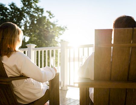 Two people sit on wooden chairs on a sunlit porch, facing a bright sunrise through a white railing.