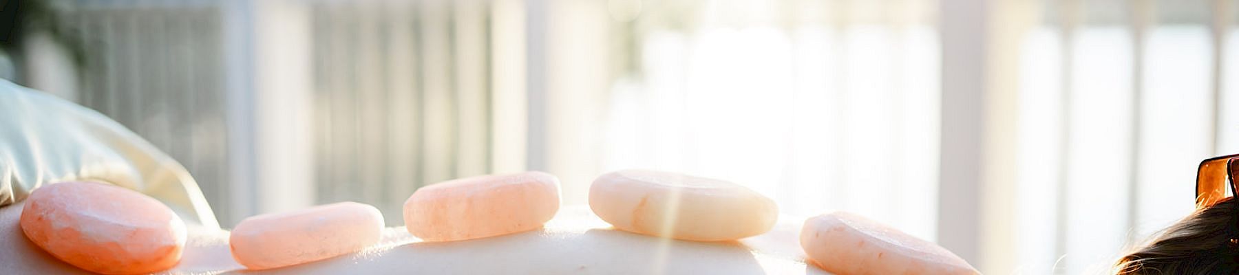 A person lying face down outdoors, with smooth stones placed along their back for a hot stone massage on a sunlit deck.