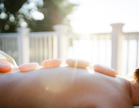 A person lying face down outdoors, with smooth stones placed along their back for a hot stone massage on a sunlit deck.