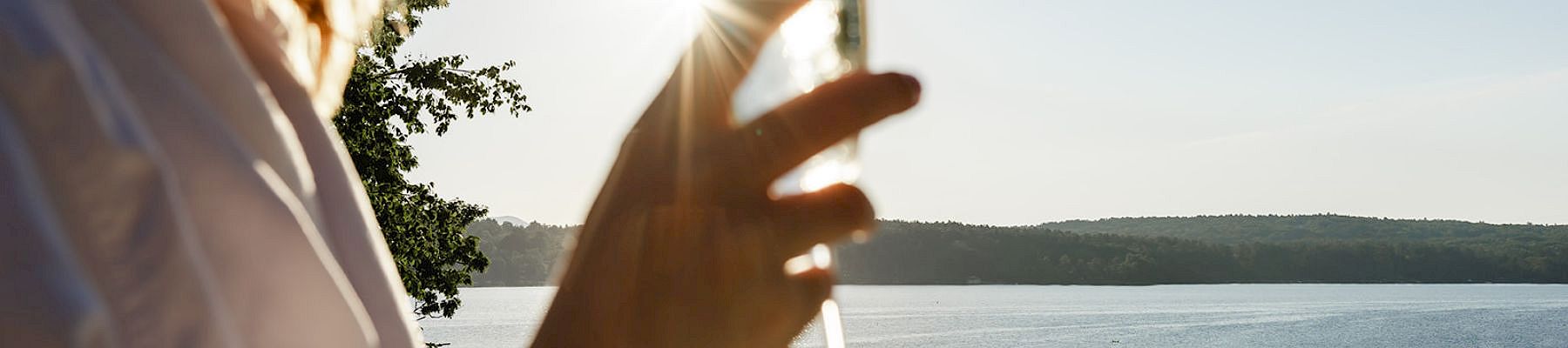 Someone holding a champagne flute by a sunny lake, smiling, with the water and distant shoreline in the background.