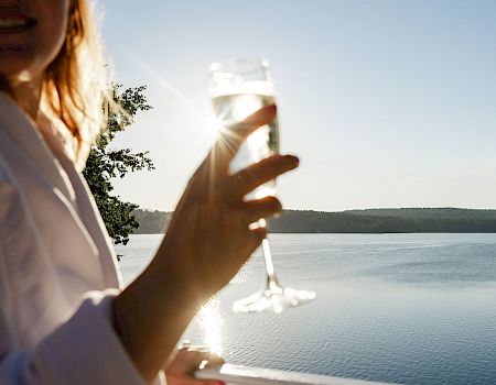 Someone holding a champagne flute by a sunny lake, smiling, with the water and distant shoreline in the background.