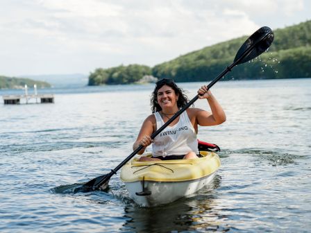 A woman paddling a yellow kayak on calm water, smiling, wearing a white tank top and life vest, with a shoreline and trees in the background. The scene is sunny.