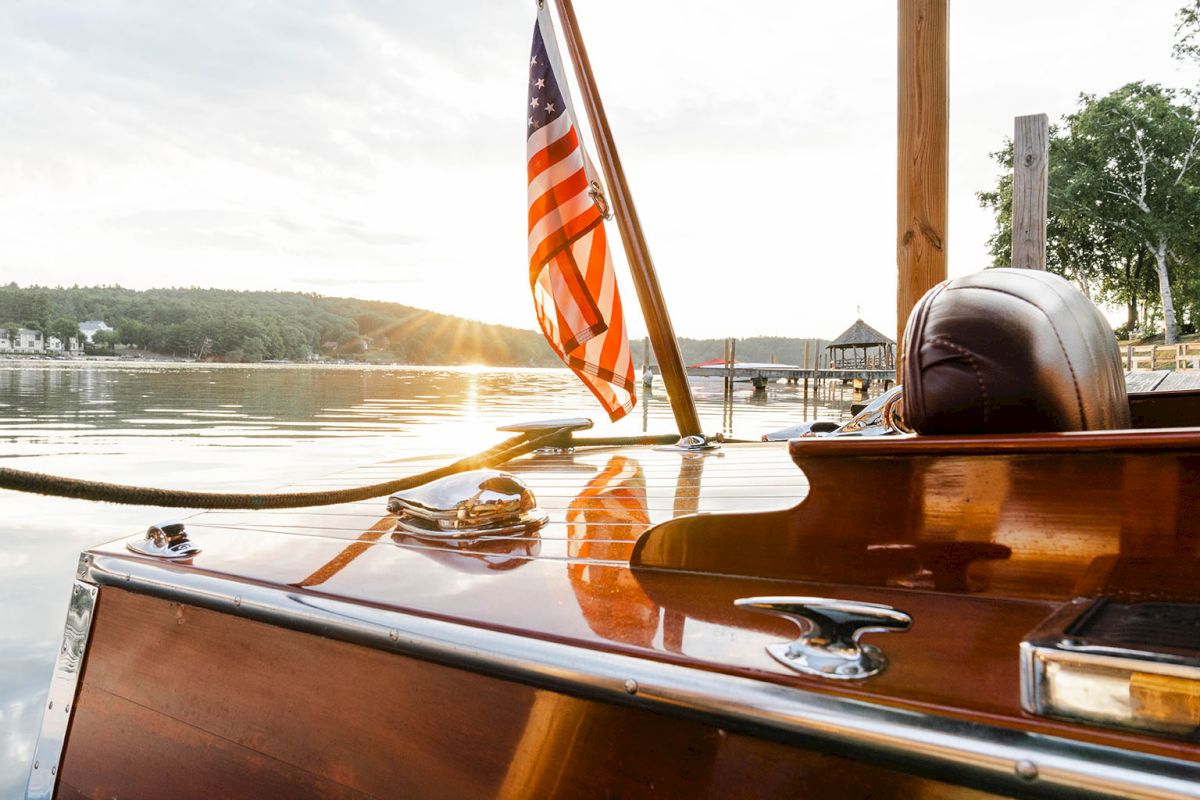 A polished wooden boat on calm water at sunset, American flag fluttering, dock and shoreline in the background.