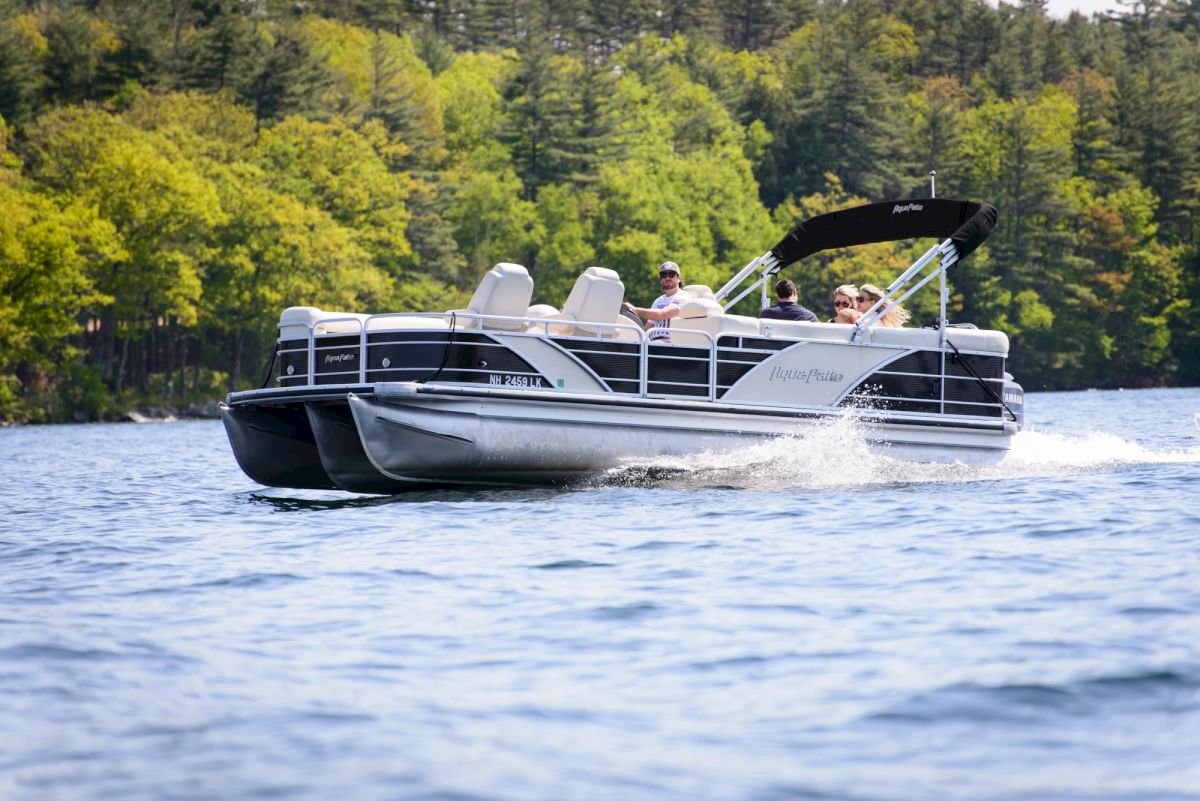 A sleek pontoon boat speeds across a lake, trees in the background, passengers enjoying a sunny ride on calm water.