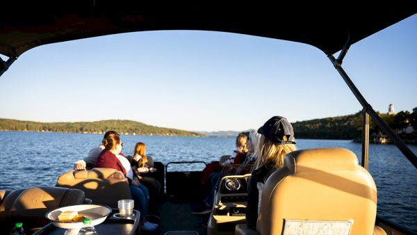 People relaxing on a boat ride, sitting in captain chairs, lake water glistening, passengers chatting as the sun sets over distant shore.