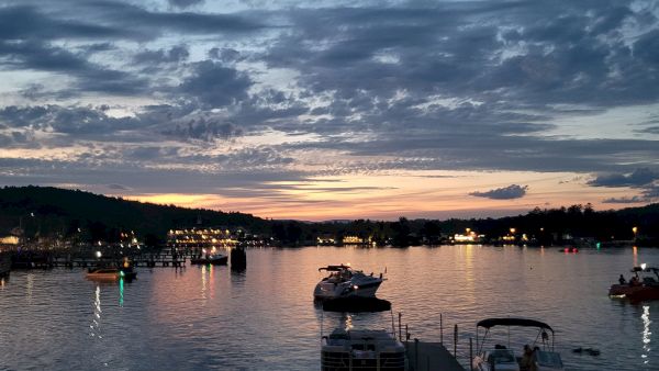 Boats docked along a calm harbor at dusk, reflecting the colorful sunset while a distant town lights up the shoreline.
