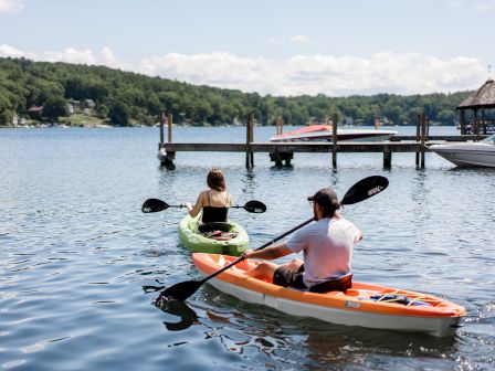 Two people kayaking on a calm lake near a wooden dock, with trees in the background and a small shaded hut on the right.