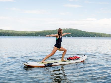 A person stands on a paddleboard, arms stretched for balance, gliding across a calm lake with distant tree-covered hills.