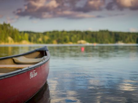 A red canoe parked at the calm lake&rsquo;s edge, reflecting a serene sky and distant tree-lined shore, with soft clouds above it.