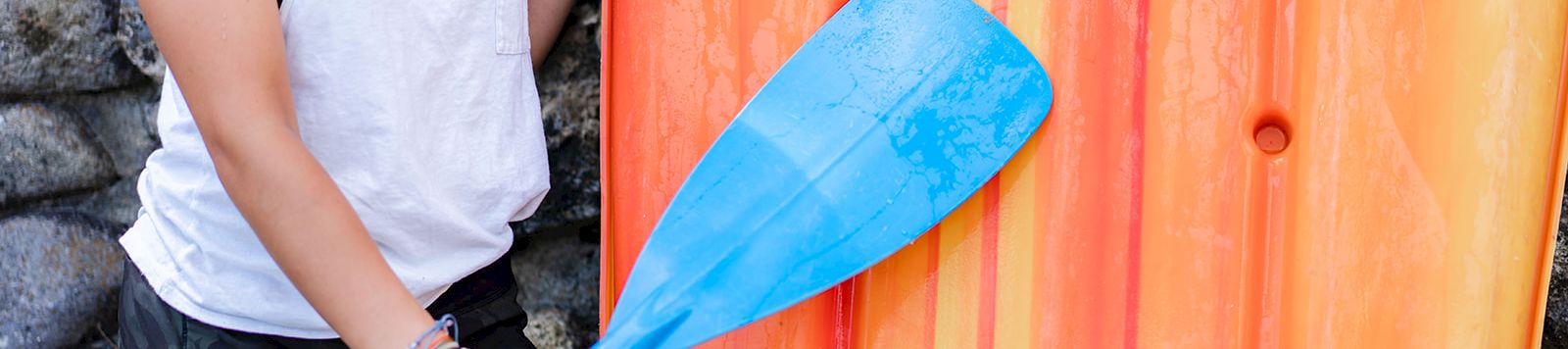 A person wearing a white tank top and black shorts holds a blue paddle next to an orange kayak. Top it at 140 characters, always ending the sentence.
