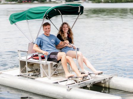 A smiling couple sits on an AquaCycle with a green canopy, calm water, and trees in the background.