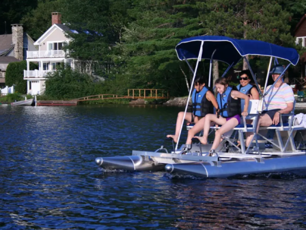 A group of people wearing life jackets sit on a 4-person AquaCycle with a canopy on a calm lake, houses and trees lining the shore.