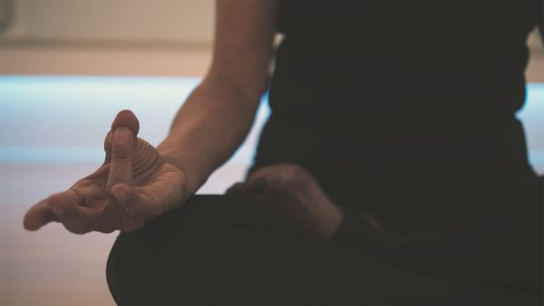 A person seated in a cross-legged yoga or meditation pose, hands resting on knees with fingers in a mudra, calm indoor setting.