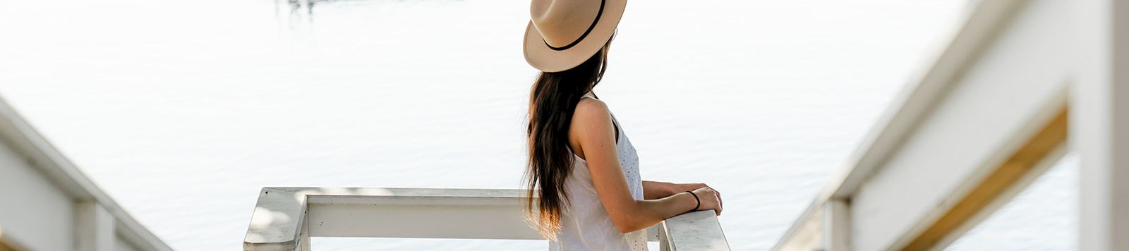 A woman stands on a white dock by the water, facing away. She wears a hat, white top, denim shorts, and gazes toward the horizon.