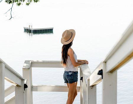 A woman stands on a white dock by the water, facing away. She wears a hat, white top, denim shorts, and gazes toward the horizon.