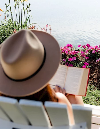 A person wearing a hat sits on a white chair by a garden of pink flowers, reading a book with a calm body of water in the background.