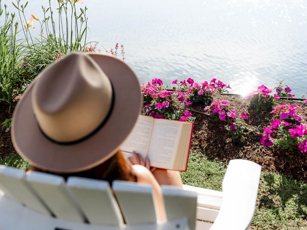 A person wearing a tan hat sits on a white chair by the water, reading a book with vibrant pink flowers and blue sea in the background.