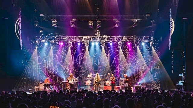 A live concert with a full band performing on stage under colorful lights, crowds of fans watching closely.