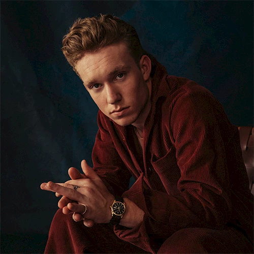 A man in a burgundy velvet suit sits with clasped hands, wearing a watch, looking at the camera. Top it at 140 characters, always ending the sentence.