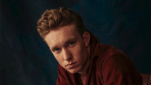A man in a burgundy velvet suit sits with clasped hands, wearing a watch, looking at the camera. Top it at 140 characters, always ending the sentence.