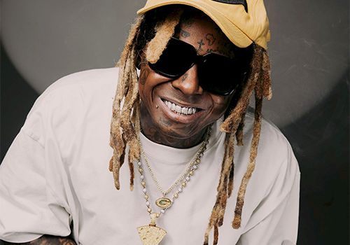A smiling man with dreadlocks and sunglasses, wearing a beige cap, white T-shirt, and gold chains.