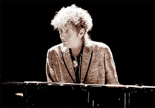 A young man with curly light hair plays a dark piano on a dim stage, wearing a patterned blazer and looking down at the keys.