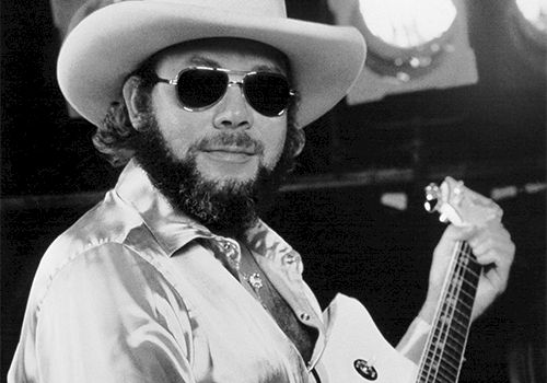 A black-and-white photo of a bearded man in a wide-brimmed hat, sunglasses, satin shirt, playing an acoustic guitar on stage.