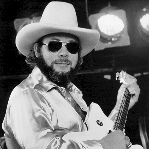 A black-and-white photo of a bearded man in a wide-brimmed hat, sunglasses, satin shirt, playing an acoustic guitar on stage.