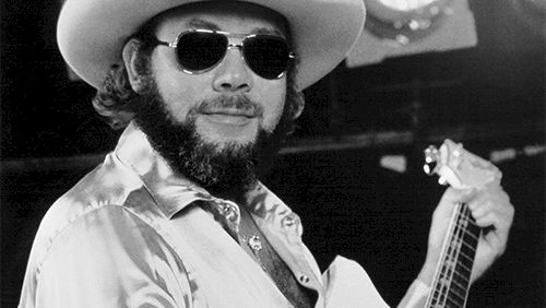 A black-and-white photo of a bearded man in a wide-brimmed hat, sunglasses, satin shirt, playing an acoustic guitar on stage.