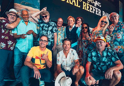 A group of casually dressed people posing together at a festival or event, tropical shirts, hat on the ground, with a "CORAL REEFER" backdrop.