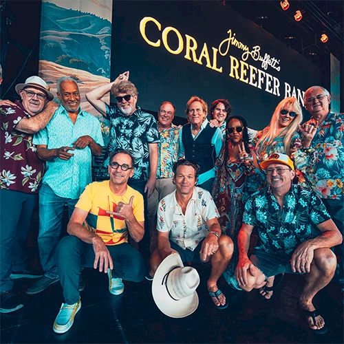 A group of casually dressed people posing together at a festival or event, tropical shirts, hat on the ground, with a "CORAL REEFER" backdrop.