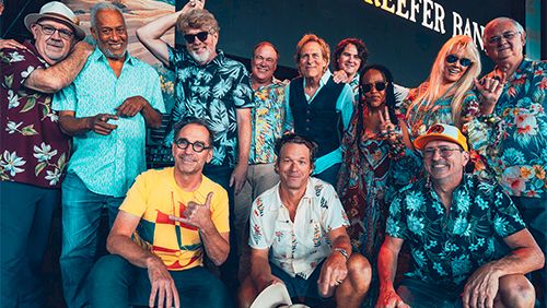 A group of casually dressed people posing together at a festival or event, tropical shirts, hat on the ground, with a "CORAL REEFER" backdrop.