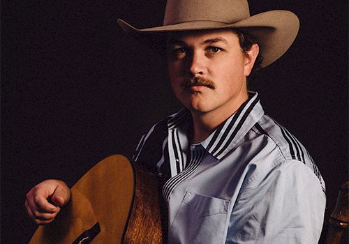 A man wearing a cowboy hat plays an acoustic guitar, looking at the camera. 140 characters, ending with a period.