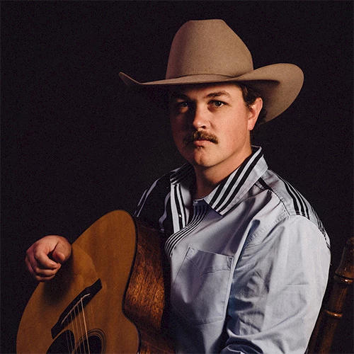 A man wearing a cowboy hat plays an acoustic guitar, looking at the camera. 140 characters, ending with a period.