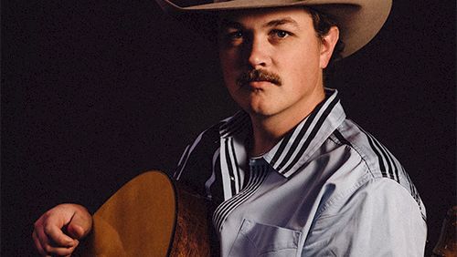 A man wearing a cowboy hat plays an acoustic guitar, looking at the camera. 140 characters, ending with a period.