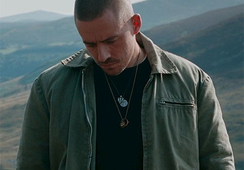 A man in a utility jacket standing outdoors on a hillside, looking down, with layered necklaces against a mountainous backdrop.