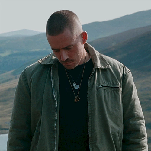 A man in a utility jacket standing outdoors on a hillside, looking down, with layered necklaces against a mountainous backdrop.