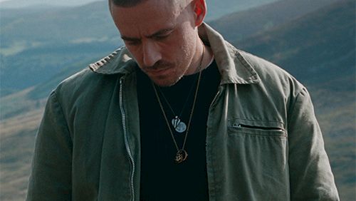 A man in a utility jacket standing outdoors on a hillside, looking down, with layered necklaces against a mountainous backdrop.