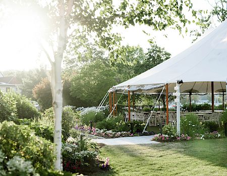 A bright outdoor garden scene with a white event tent, greenery, flowering beds, and sunlight streaming through trees, creating a cheerful setup.