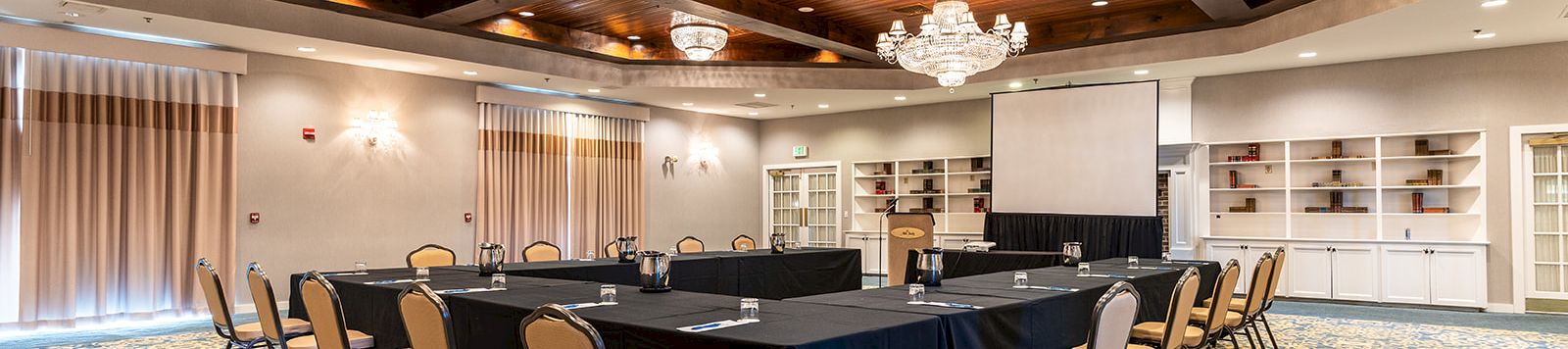 A conference room with a long U-shaped table, black tablecloths, beige chairs, crystal chandeliers, wood-paneled ceiling, and a patterned blue carpet.