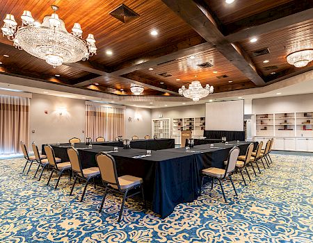 A conference room with a long U-shaped table, black tablecloths, beige chairs, crystal chandeliers, wood-paneled ceiling, and a patterned blue carpet.