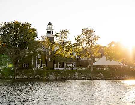 A lakeside town with trees, a brick building and clock tower along the shore, bathed in warm sunset light on calm water.