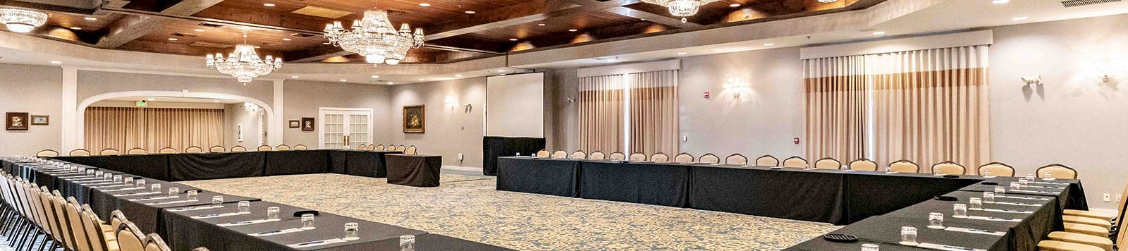 A large conference room with U-shaped black-table setup, beige chairs, chandeliers, carpeted floor, and nameplates/water glasses at each seat.