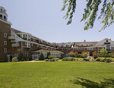 A large, brown multi-story hotel with a tower on the left, white railings, and a manicured green lawn in the foreground, sunny day.