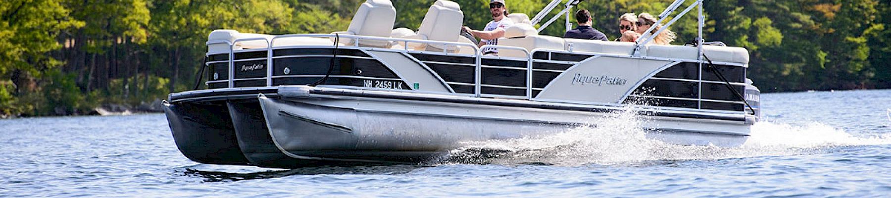A sleek pontoon boat glides across calm water near a tree-lined shoreline, two passengers enjoying the ride as the wake trails behind the craft.