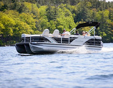 A sleek pontoon boat glides across calm water near a tree-lined shoreline, two passengers enjoying the ride as the wake trails behind the craft.
