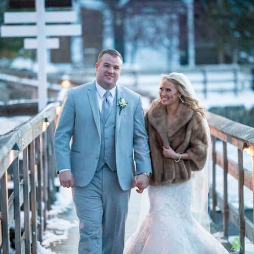 A newlywed couple walks hand-in-hand on a snowy bridge; he in a light gray suit, she in a white gown with a fur wrap.