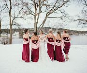 Six brides wearing matching magenta gowns stand in a snowy park, backs to camera, arms around each other, trees and a lake in the background.