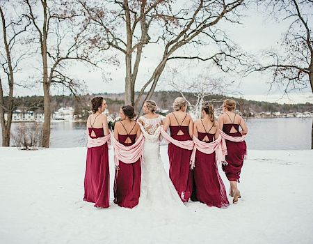 Six brides wearing matching magenta gowns stand in a snowy park, backs to camera, arms around each other, trees and a lake in the background.